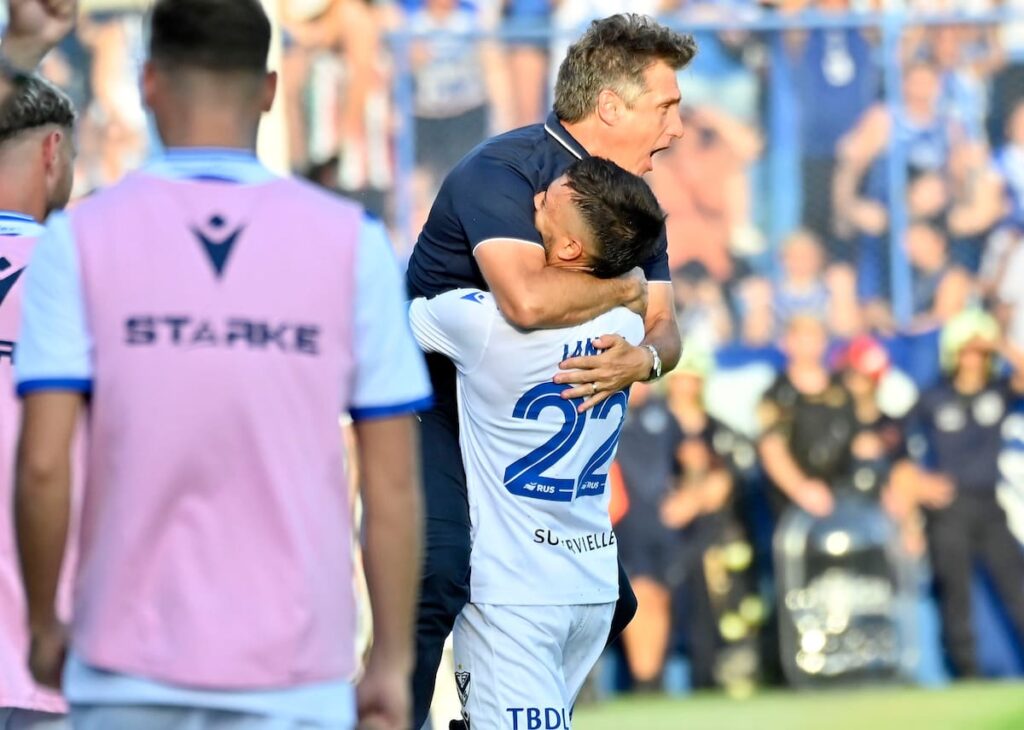 Carlos Tevez de espaldas a Guillermo Barros Schelotto en el banco de Vélez, con mirada tensa y el campo de Liniers al fondo