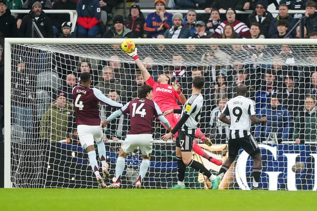 Dibu Martínez en acción con guantes amarillos deteniendo un remate en el St. James Park durante el Newcastle vs Aston Villa
