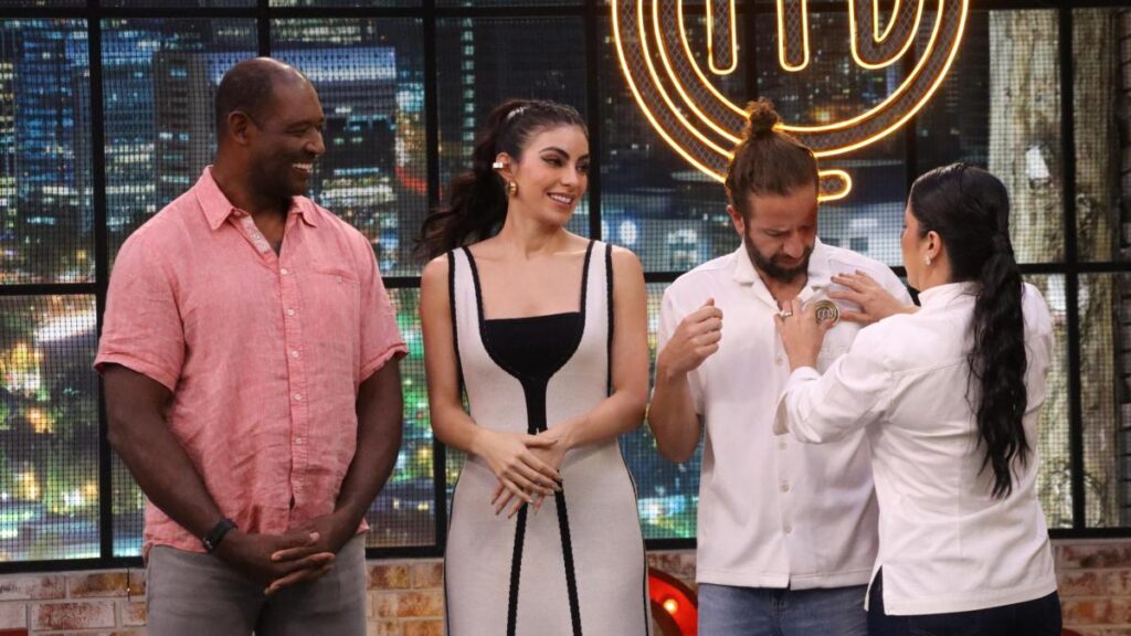 Bastián Napolitano sostiene el pin del chef en MasterChef con expresión decidida frente a Beber Espinoza