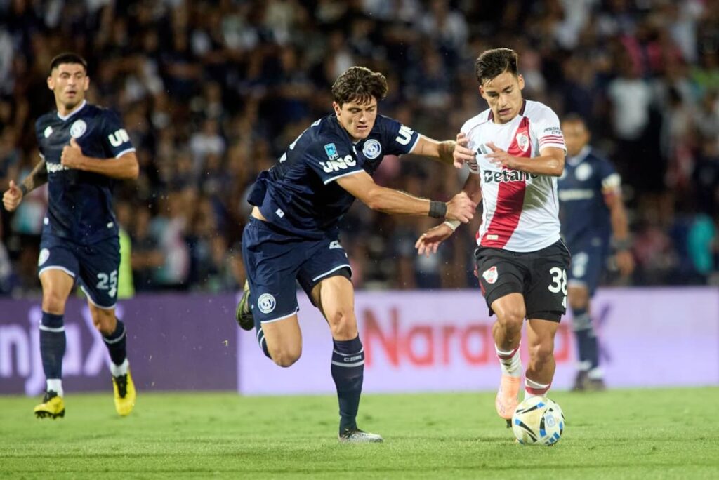 Eduardo Coudet analiza tácticas en un banco vacío mientras River sufre en defensa, con Beltrán atajando un remate de cabeza