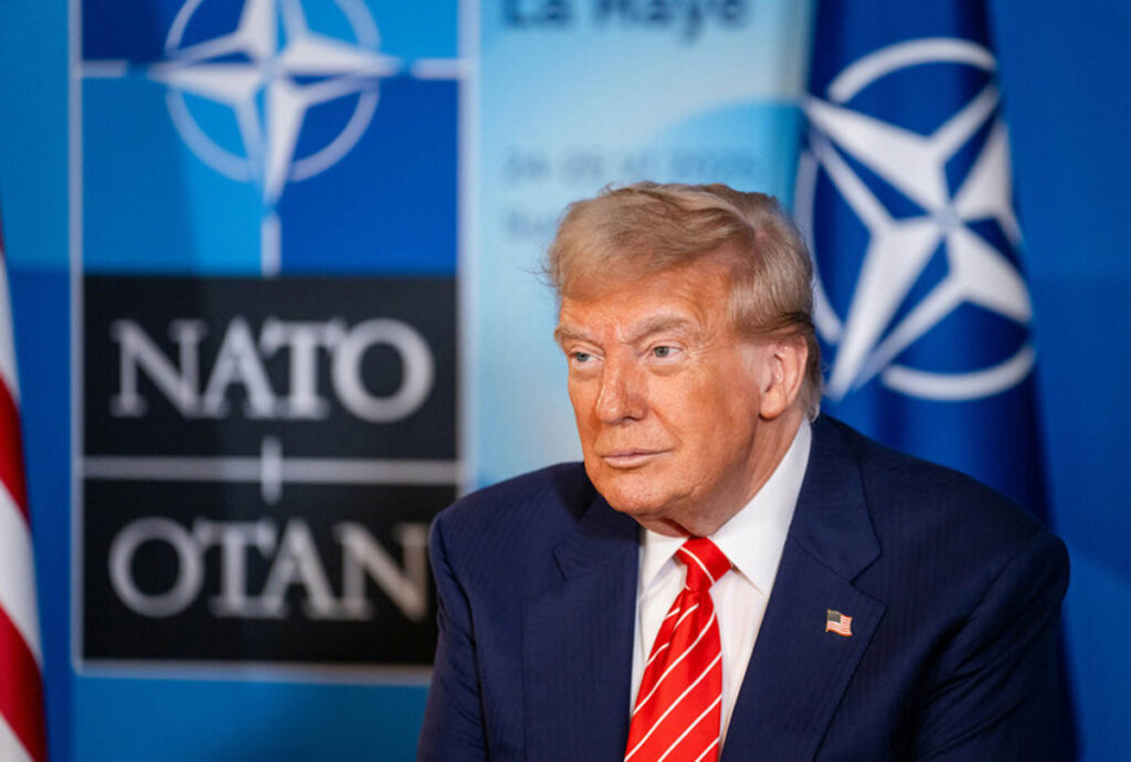 Donald Trump en mitin señalando con gesto serio mientras un cartel de la OTAN aparece difuminado al fondo