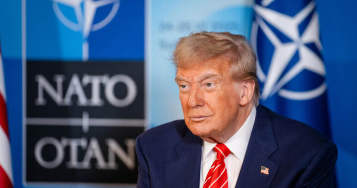 Donald Trump en mitin señalando con gesto serio mientras un cartel de la OTAN aparece difuminado al fondo
