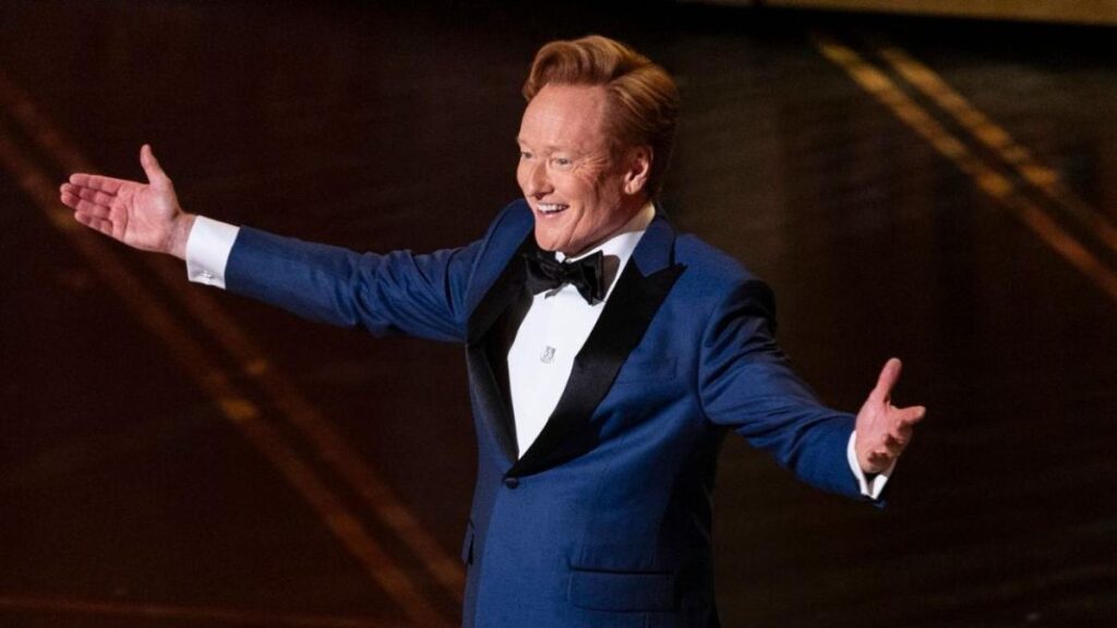 Conan O'Brien sonríe en el escenario de los Oscar 2026 con fondo del Dolby Theatre, listo para su histórico tercer año como anfitrión en 2027