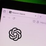 Logotipos de OpenAI y Anthropic en pantalla de teléfono móvil con fondo de bolsa de valores